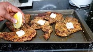 Person squeezing a lemon onto pork chops topped with butter.