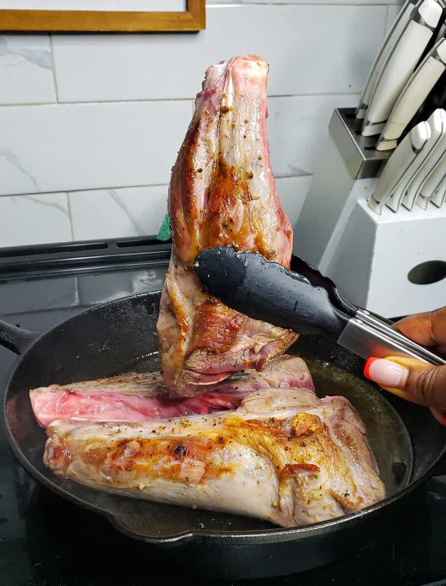 Tongs flipping a seared lamb shank.