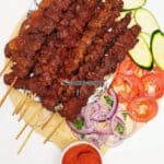 Beef Suya on a wooden board with sliced vegetables.
