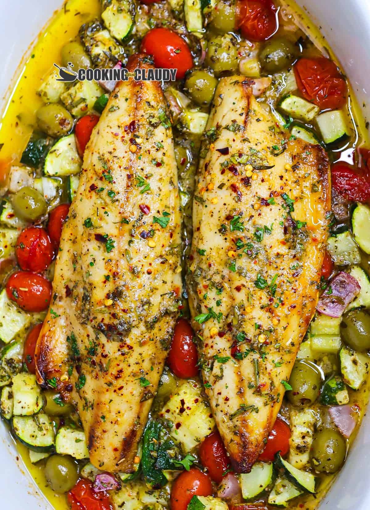 Mediterranean baked fish on top veggies in a white baking dish.