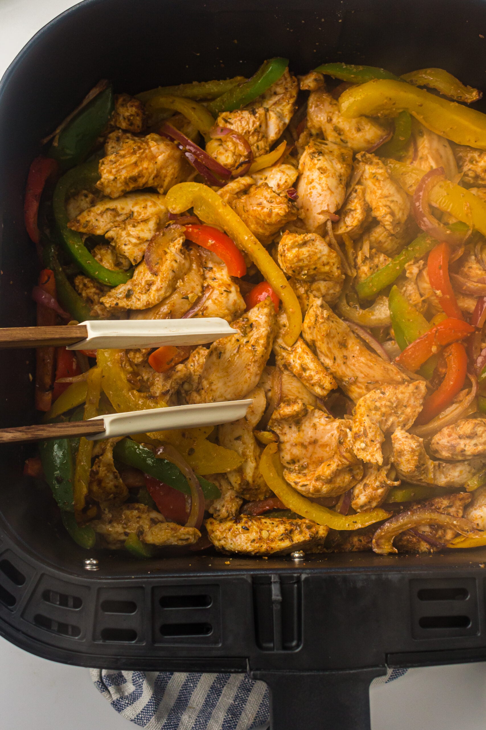 Tongs turning chicken fajita strips in the air fryer basket
